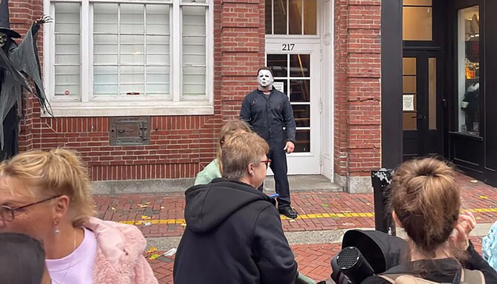 A person wearing a ghostly white mask stands behind a group of unaware pedestrians on a city sidewalk adding a spooky element to the scene