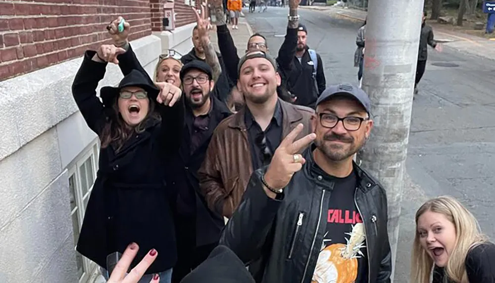 A group of cheerful people are posing for a photo on a city street some of them making peace signs or pointing towards the camera