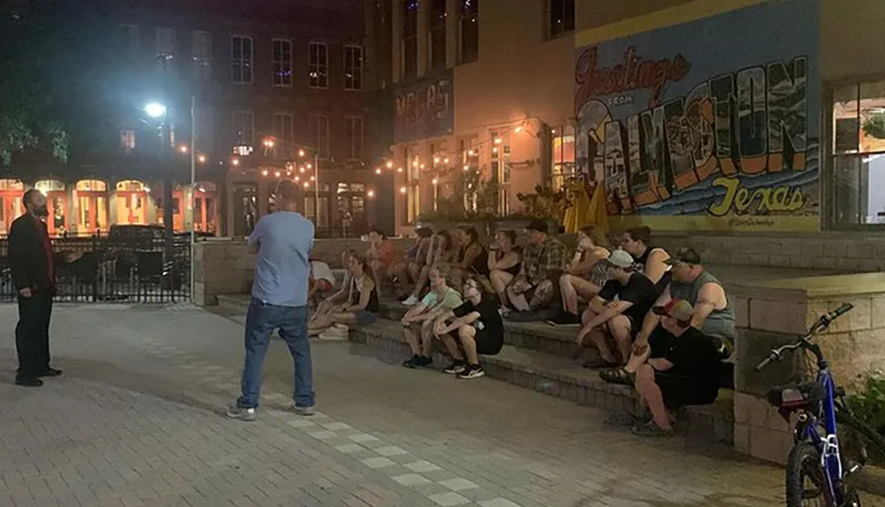 A group of people is sitting outdoors on steps at night listening to a man standing opposite them with a colorful mural wall in the background