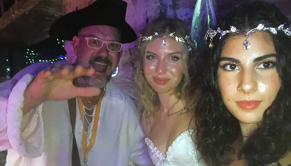 A man in a pirate hat extends his hand toward the camera while posing with two women wearing festive headpieces at an apparent party or costume event