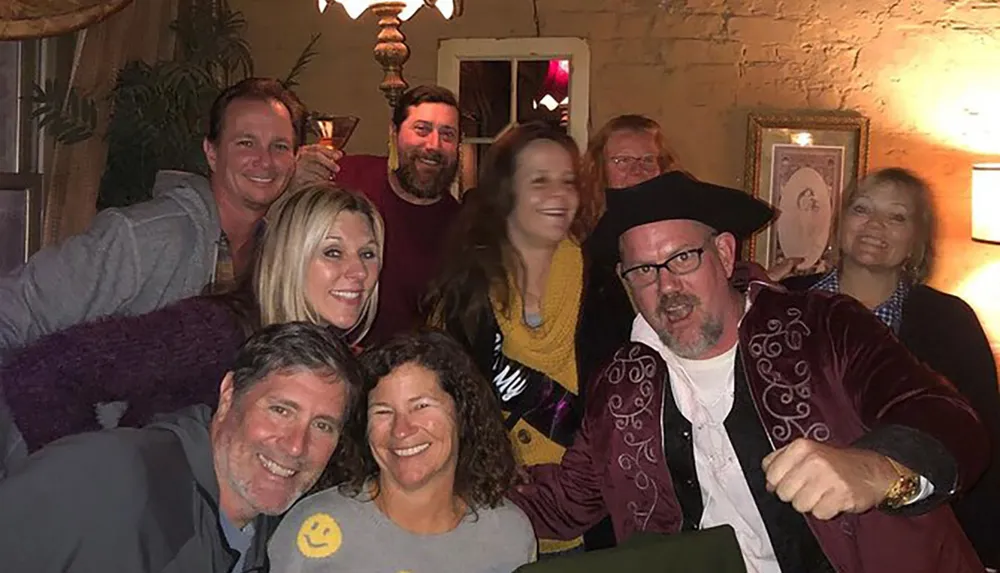 A group of smiling people are posing for a photo in a warmly lit room with one person in the foreground wearing a playful pirate hat