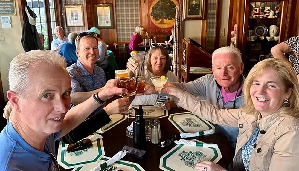 A group of five smiling adults are toasting with drinks at a table inside a restaurant with traditional decor