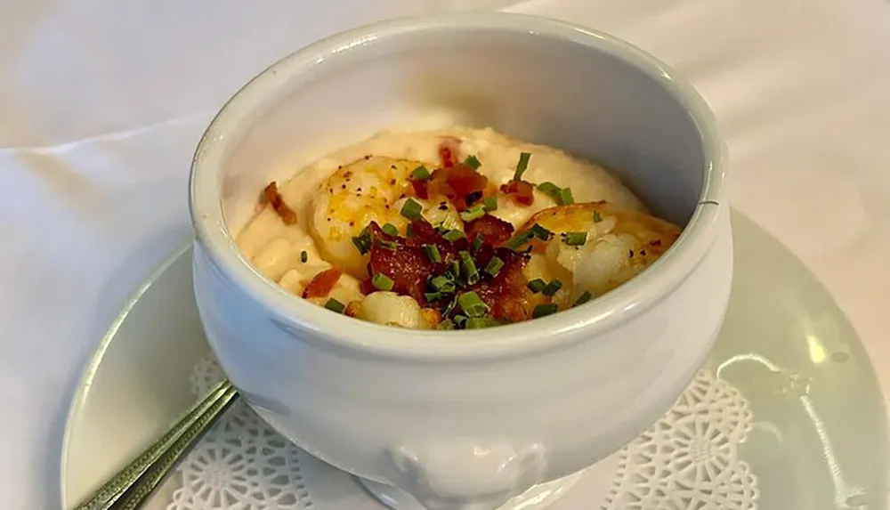 The image shows a serving of shrimp and grits garnished with bacon bits and chives in a white bowl placed on a white plate with a decorative white paper doily under the bowl