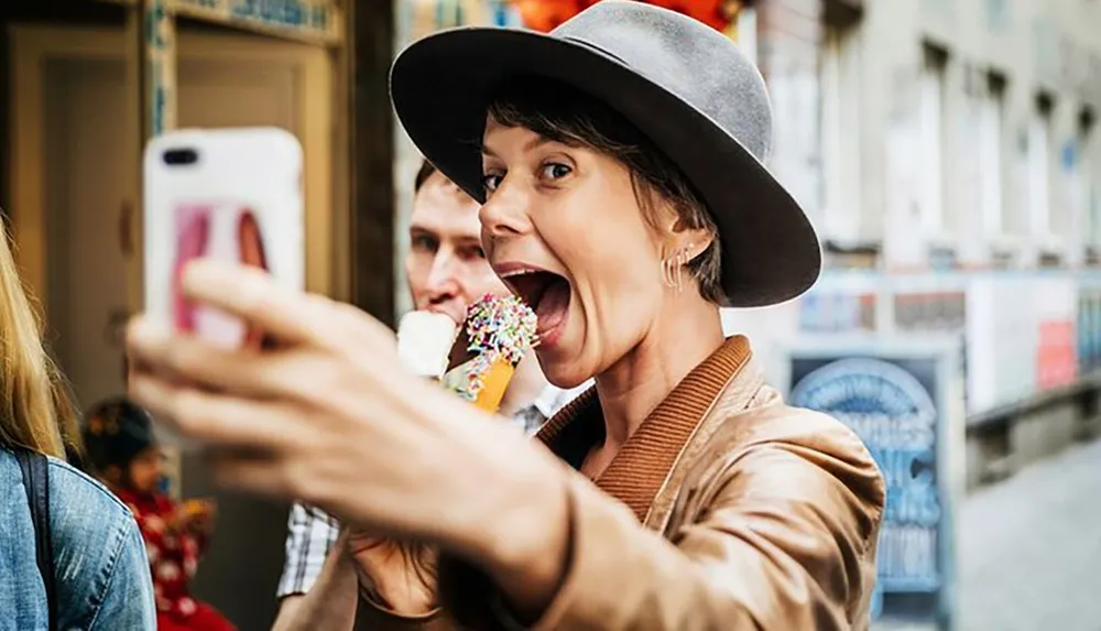 A person wearing a gray hat excitedly poses for a selfie while taking a bite of an ice cream cone with a slightly out-of-focus man in the background