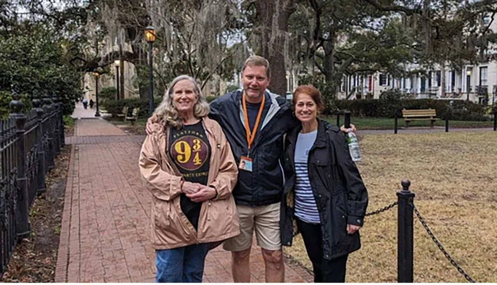 Three people are posing for a photo on a tree-lined path with a wrought iron fence on the left and a park bench in the background