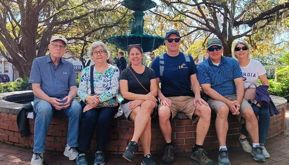 Six people are smiling and sitting on a bench in front of a fountain in a park with abundant greenery and sunshine