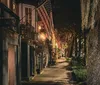 The image displays a charming nocturnal street view adorned with American flags hanging from traditional buildings illuminated by warm streetlights and lined with trees draped in Spanish moss