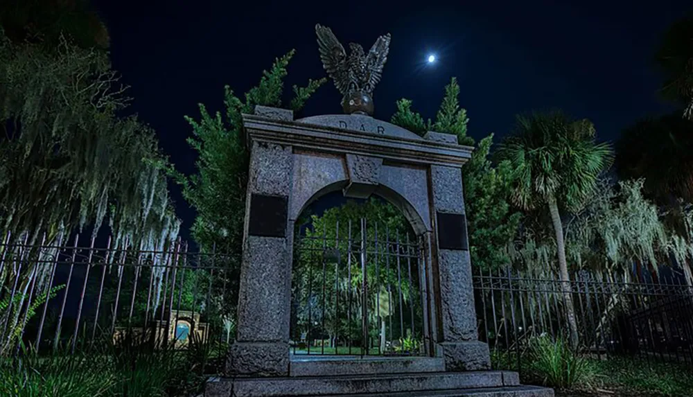 The image depicts a nighttime scene of an ornate gate with an eagle sculpture atop surrounded by Spanish moss-draped trees under a moonlit sky