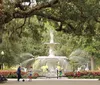 The image depicts a serene park setting with people walking near an ornate fountain surrounded by lush greenery and hanging Spanish moss from the trees