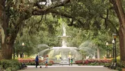 The image depicts a serene park setting with people walking near an ornate fountain, surrounded by lush greenery and hanging Spanish moss from the trees.