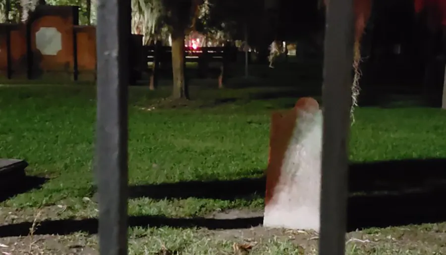 The image is a nighttime scene showing a grassy area viewed between vertical metal bars, with possibly a headstone or a monument in the middle ground and trees in the background.