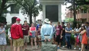 A group of people appear to be on an outdoor guided tour, listening to a person who is likely the tour guide standing in front of them with his back to the camera.