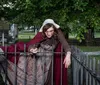 A person in historical costume is leaning over an iron fence in a cemetery portraying a dramatic expression
