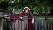 A person in historical costume is leaning over an iron fence in a cemetery, portraying a dramatic expression.