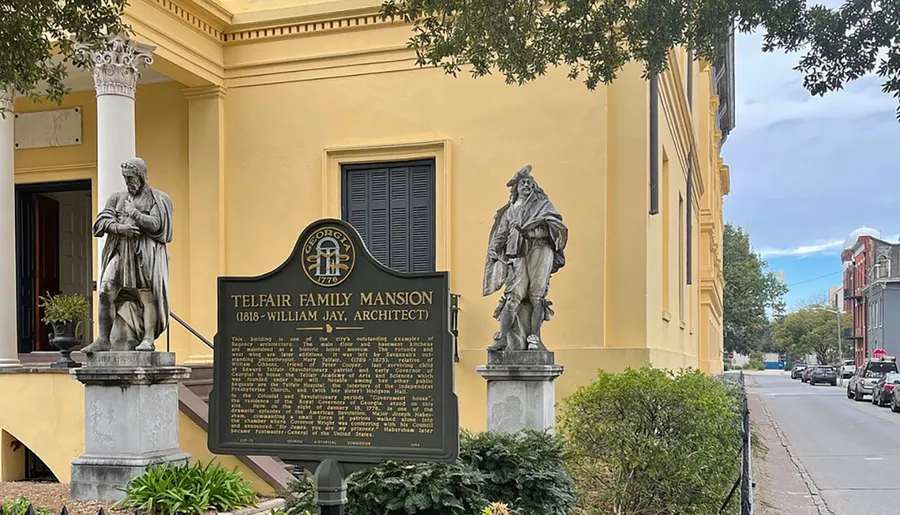 The image shows two statues flanking a historical marker in front of the yellow Telfair Family Mansion, with an urban street scene in the background.