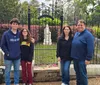 A family of four is posing for a photo in front of an ornate metal gate that leads to a cemetery with mature trees and tombstones in the background