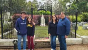 A family of four is posing for a photo in front of an ornate metal gate that leads to a cemetery, with mature trees and tombstones in the background.