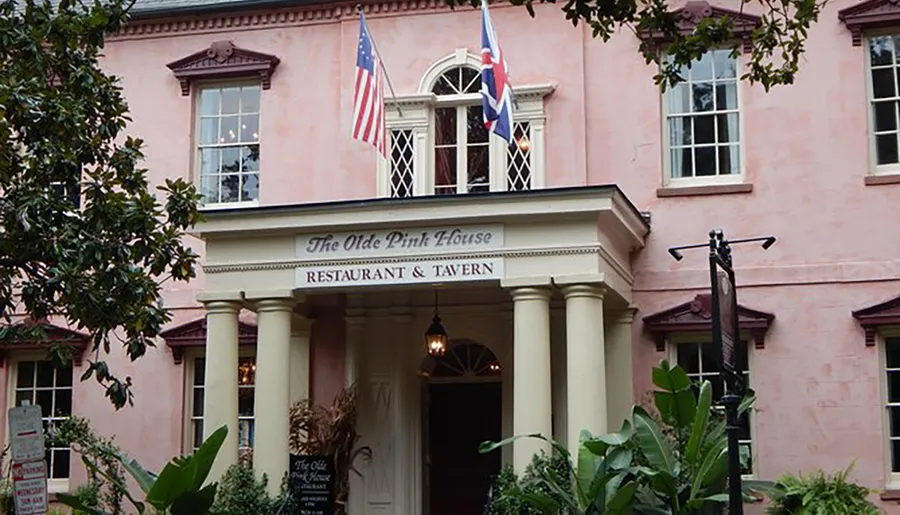 The image shows the front facade of The Olde Pink House, a historic restaurant and tavern, with its signature pink exterior and colonial architectural elements, flanked by American and state flags under a cloudy sky.