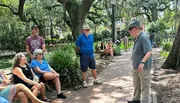 A group of people listens to a speaker in a park with lush trees and Spanish moss, while others enjoy the surroundings.