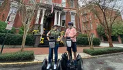 Two people are smiling for the camera while standing on Segway scooters in front of a building with a historic facade.
