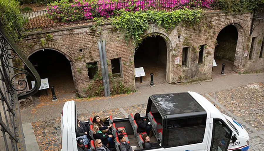 A tour bus with passengers is passing beneath a stone bridge adorned with flowering plants in an historic setting.