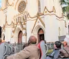 A group of tourists enjoys a guided tour in an open-top Kelly Tours vehicle passing by a park with lush trees and historic architecture