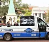 A group of tourists enjoys a guided tour in an open-top Kelly Tours vehicle passing by a park with lush trees and historic architecture