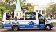 A group of tourists enjoys a guided tour in an open-top Kelly Tours vehicle passing by a park with lush trees and historic architecture.