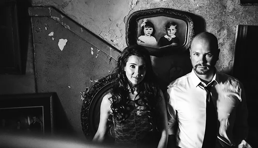 A black and white photo featuring a woman and a man posing solemnly in a room with peeling walls and vintage framed photos