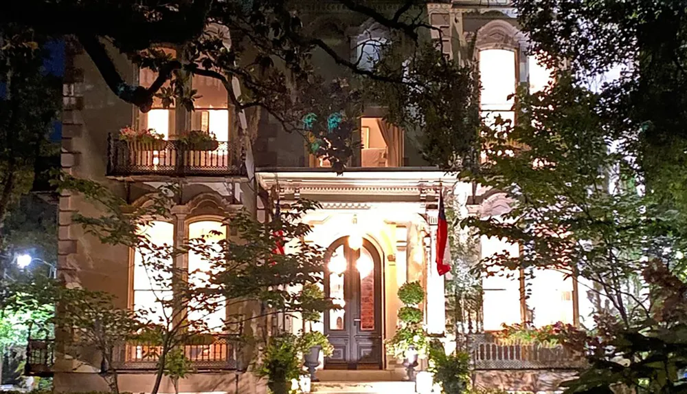 The image shows an illuminated ornate building at night partially obscured by trees with warm lighting shining from the windows and a flag hanging on the right side