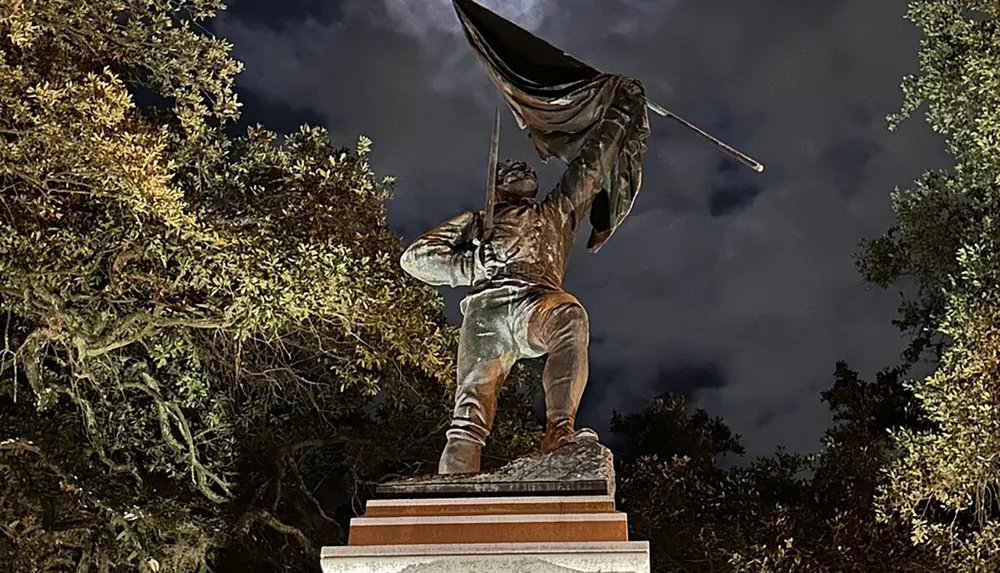 The image shows a bronze statue of a figure holding a flag aloft against a night sky with trees in the background