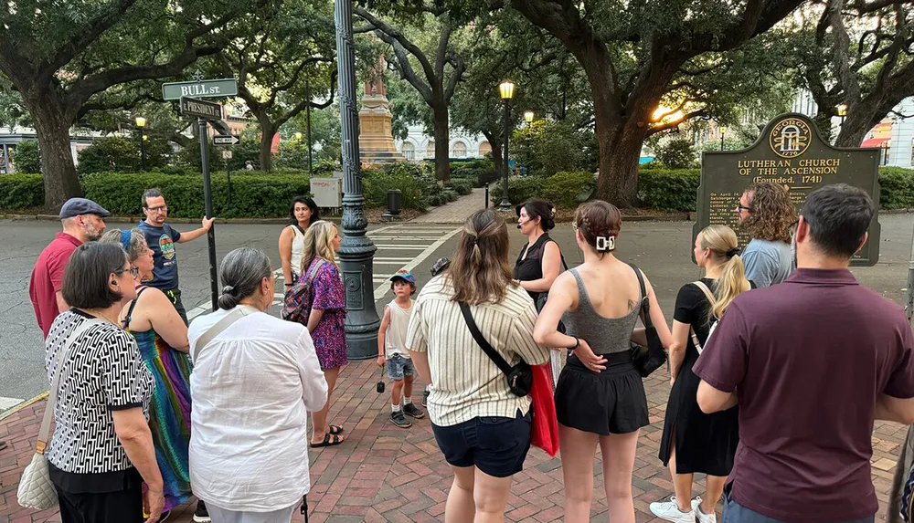 A group of people are attentively listening to a man who appears to be giving a tour at an urban street corner near a sign for the Lutheran Church of the Ascension