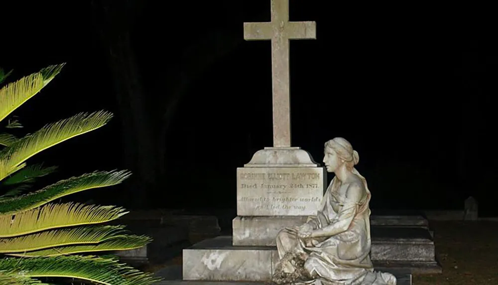 A statue of a seated figure gazing at a cross which forms part of a tombstone is captured at night with illuminated foliage nearby