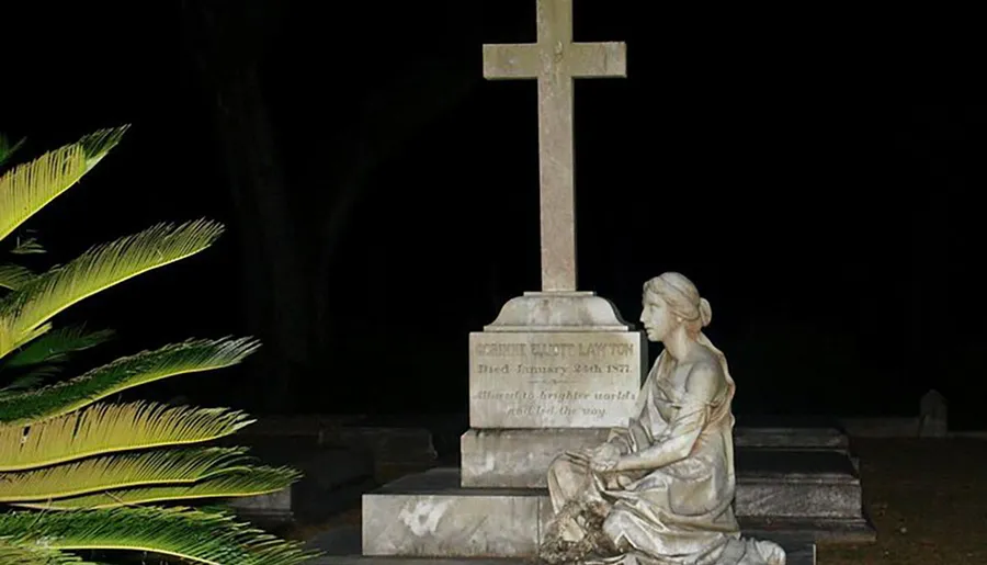 A statue of a seated figure gazing at a cross, which forms part of a tombstone, is captured at night with illuminated foliage nearby.