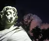 The image shows a white statue of a young girl in vintage clothing illuminated at night with a dark hedge in the background