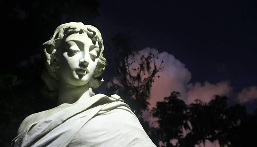 A statue of a woman is illuminated from the side against a dramatic evening sky with clouds and trees in the background