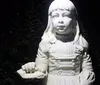 The image shows a white statue of a young girl in vintage clothing illuminated at night with a dark hedge in the background