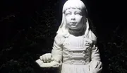 The image shows a white statue of a young girl in vintage clothing, illuminated at night with a dark hedge in the background.