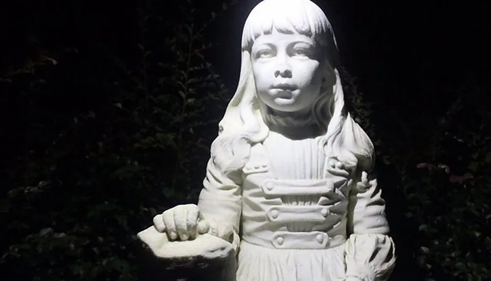 The image shows a white statue of a young girl in vintage clothing illuminated at night with a dark hedge in the background