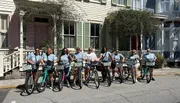 A group of smiling people are standing with rented bicycles in front of a house with a charming facade.