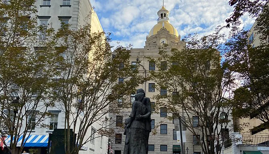 A statue of a standing figure is seen in the foreground against a backdrop of trees and a historic building with a gold-domed tower.