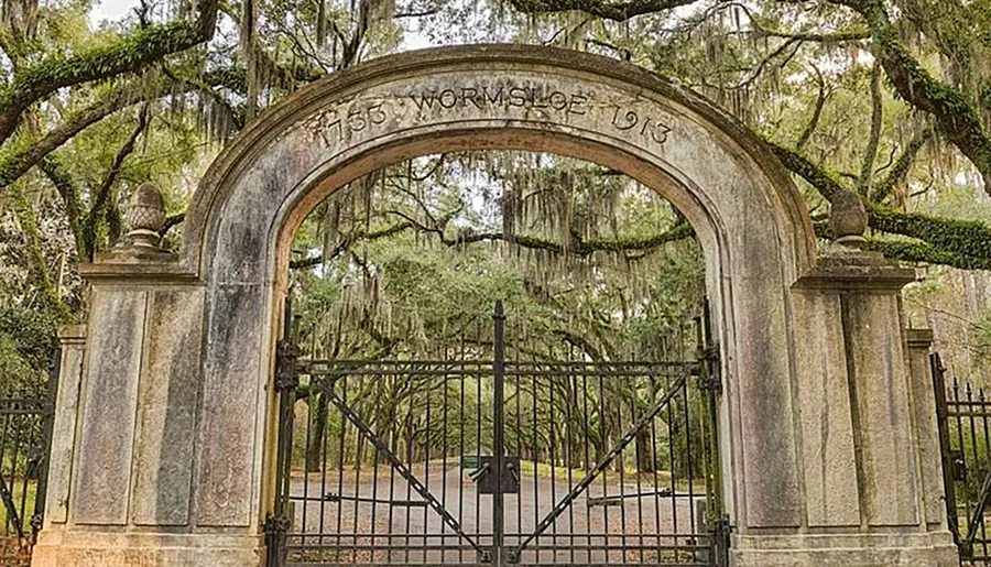The image shows an ornate, historical stone gate with the inscribed words 1750 WORMSLOE 1913, flanked by hanging Spanish moss and a metal fence, suggestive of being an entrance to an estate or historical site.