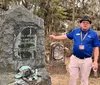 A person gives a presentation or tour in a cemetery pointing at a large rugged gravestone with detailed engravings and metal adornments