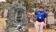 A person gives a presentation or tour in a cemetery, pointing at a large, rugged gravestone with detailed engravings and metal adornments.