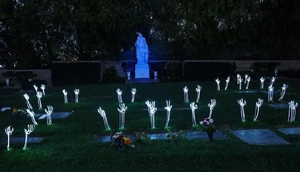 The image depicts a night scene in a cemetery with multiple glow-in-the-dark skeleton hands protruding from the ground centered around a statue illuminated in blue light