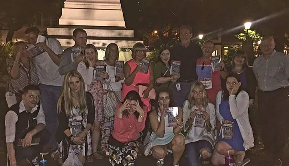 A group of people are posing with various expressions and hand gestures some holding what appears to be a book or pamphlet at an outdoor nighttime gathering