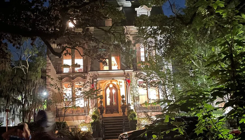 The image is a nighttime view of a grand multi-story house with lit windows a front staircase and festive decorations partially obscured by trees in a setting that appears both eerie and alluring