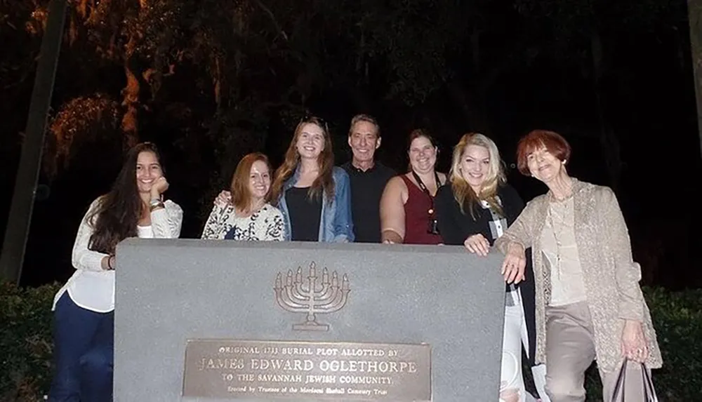 A group of seven people is smiling for a photo at night in front of a historical marker commemorating the original 1733 burial plot allotted by James Edward Oglethorpe to the Savannah Jewish community