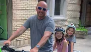A man with two smiling children wearing helmets are preparing for a bike ride.