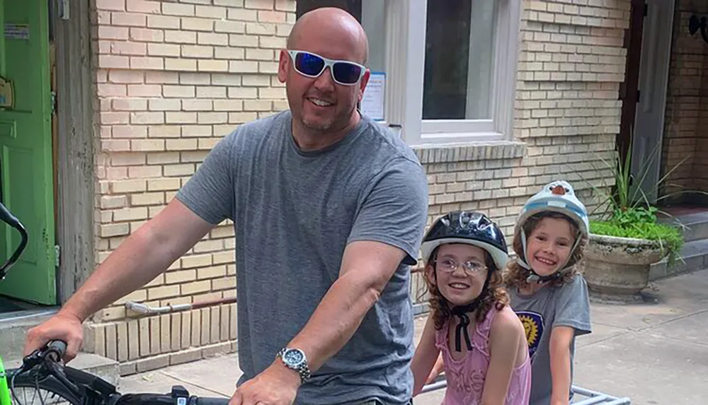 A man with two smiling children wearing helmets are preparing for a bike ride